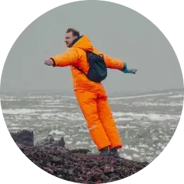 Aufgeregter Reisender, der die isländische Landschaft umarmt Mann in leuchtend orangefarbener Winterkleidung, der auf felsigem Terrain mit ausgebreiteten Armen steht.