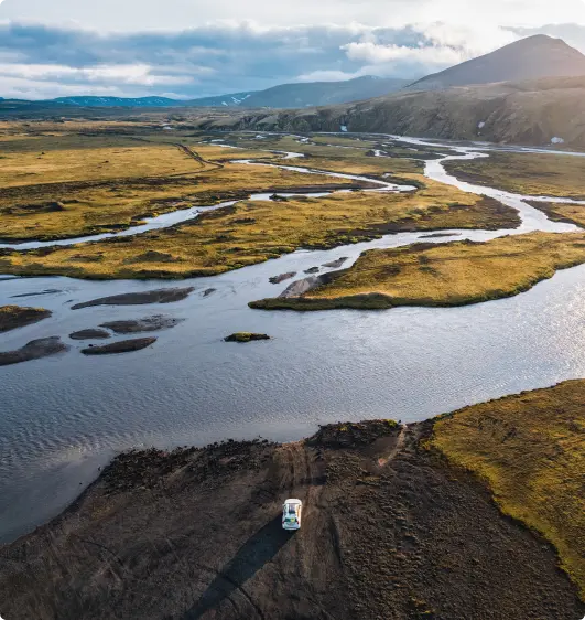 Ein einsamer 4x4-SUV parkt in der weiten, offenen Wildnis Islands, umgeben von gewundenen Wasserläufen, grasbewachsenen Ebenen und fernen Bergen unter einem bewölkten Himmel.