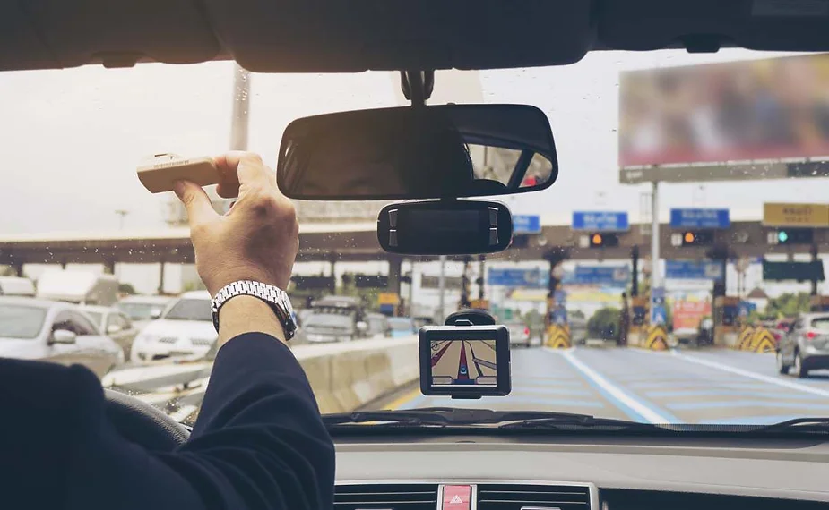 Ein Fahrer im Auto greift mit einem Maut-Transponder zum Bereich an der Windschutzscheibe, um in eine Mautspur einzufahren; auf dem Armaturenbrett zeigt ein GPS die Navigation, und voraus sind weitere Fahrzeuge zu sehen.