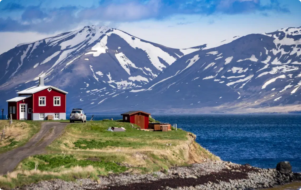 Ein idyllisches isländisches Haus am Wasser mit schneebedeckten Bergen im Hintergrund ist der perfekte Ausgangspunkt für eine Rundreise durch Island. Am internationalen Flughafen Keflavik können Sie einen Mietwagen für Ihre Reise abholen, mit einer Auswahl an Personenkraftwagen, Elektroautos und Luxusmodellen für ein unvergessliches Abenteuer.