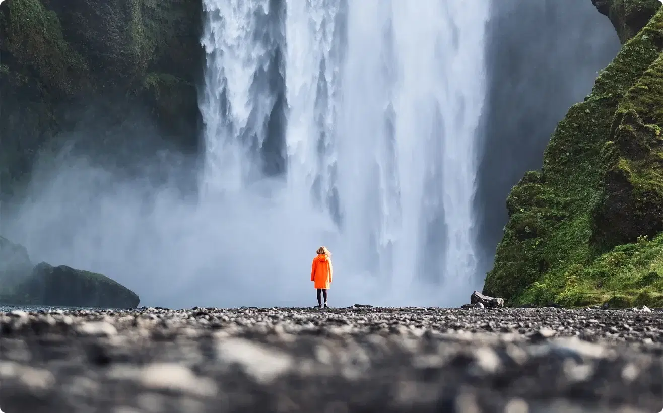 Reisender in oranger Jacke vor dem Skógafoss – Sinnbild unserer Mission, unvergessliche Erlebnisse zu schaffen.